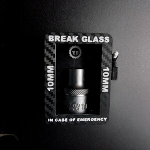 May include: A black and grey tool kit with the text "BREAK GLASS" and "IN CASE OF EMERGENCY". The kit contains a 10mm socket. The socket is silver and has a textured grip. The kit is on a black surface.