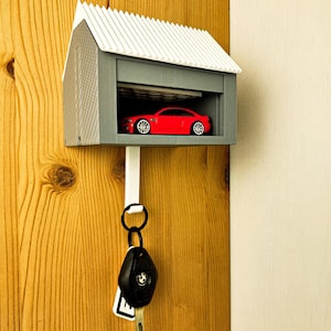 Op de afbeelding: Een sleutelhouder in de vorm van een miniatuur garage. De grijze structuur heeft een wit dak en een rode speelgoedauto binnenin. Een sleutelring met sleutels hangt eronder. De sleutelhouder is bevestigd op een houten deur.