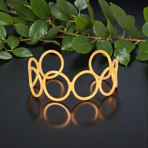 May include: Gold-toned open cuff bracelet with a design of linked circles. The bracelet is set against a dark backdrop with green foliage, accentuating the jewellery's form and colour. The bracelet's design is modern.