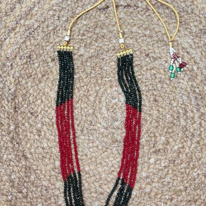 May include: A multi-strand necklace featuring alternating sections of red and dark green beads. The necklace has a gold-colored chain and a small decorative charm with red and green accents. The beads are strung together to create a layered effect.