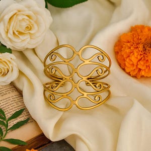 May include: Gold-toned metal cuff bracelet with an openwork design. The bracelet features a series of interconnected, symmetrical swirls. The jewelry is displayed on a cream-colored fabric, with white roses and an orange flower in the background.
