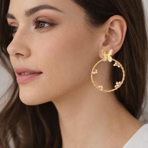 May include: Gold-coloured hoop earrings with a butterfly detail at the top and small pearl-like beads. The hoops have a twisted rope design. The earrings are worn on a person's ear.
