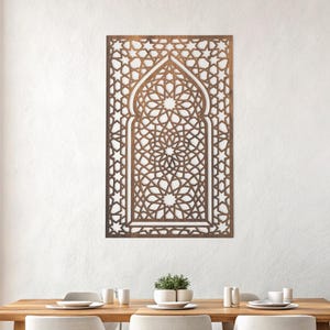 Puede incluir: Una pieza de arte mural de madera marrón con un intrincado patrón geométrico, que se asemeja a una ventana o puerta arqueada. El diseño presenta estrellas y otras formas, creando un elemento decorativo y artístico. El arte está montado en una pared blanca sobre una mesa de comedor.
