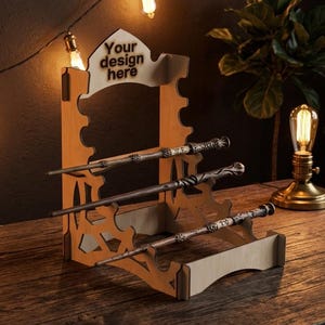May include: A light-colored wooden display stand with three horizontal tiers designed to hold wands. The top section features a cutout area with the text "Your design here" printed in dark letters. The stand has a laser-cut, open-work side profile.