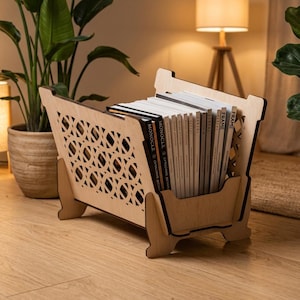 May include: A light-colored wooden magazine rack featuring intricate laser-cut circular patterns on the side panels. The rack is filled with a collection of magazines, including titles such as Monocle and Frame, arranged vertically in the center.