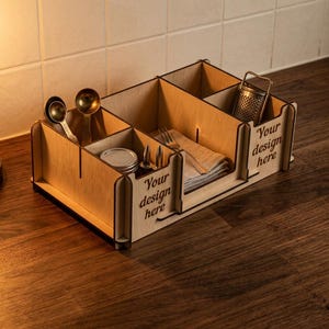 Könnte beinhalten: Ein Küchenorganizer aus hellem Holz mit mehreren Fächern für Besteck und Servietten. Die Seitenteile sind mit dem lasergravierten Text Your design here versehen. Der Organizer enthält zwei Messlöffel, ein kleines Glas und eine Metallreibe.