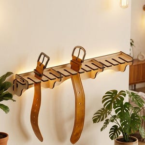 Può includere: Un portacinture da parete in legno chiaro dotato di molteplici fessure per appendere le cinture. Il supporto include una retroilluminazione integrata e sostiene due cinture in pelle color cuoio con fibbie in metallo dal design funzionale.