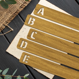 May include: Five wooden bookmarks, each with a letter from A to E cut out. The bookmarks are a warm, golden brown, with a wood grain texture. They are arranged on off-white paper, set against a black wooden surface in the background.