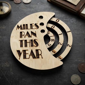 Könnte beinhalten: Ein halbmondförmiger Tracker aus hellem Holz mit der eingravierten Aufschrift MILES RAN THIS YEAR. Er verfügt über drei konzentrische, drehbare Ringe mit den Zahlen null bis neun zur manuellen Einstellung der Kilometerstände.