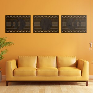 Könnte beinhalten: Drei Wandkunst-Paneele aus dunklem Holz, die Mondphasen darstellen. Jedes Paneel hat eine rechteckige Form mit einem geometrischen Design. Die Paneele sind über einem gelben Sofa an einer orangefarbenen Wand angebracht.
