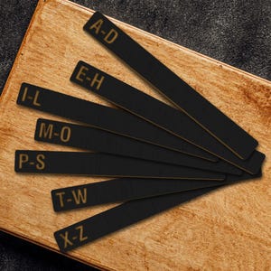 May include: Black rectangular wooden bookmarks with gold lettering. Each bookmark displays a range of letters: A-D, E-H, I-L, M-O, P-S, T-W, and X-Z. The bookmarks are arranged on a wooden surface.
