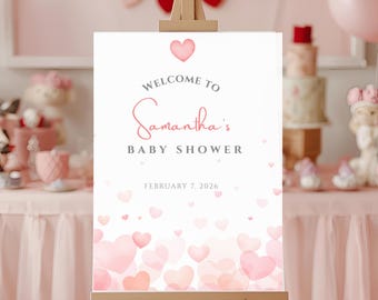 May include: A baby shower welcome sign on a wooden easel. The sign is white with pink hearts and text that reads "Welcome to Samantha's Baby Shower" and the date February 7, 2026. Pink heart balloons and decorations are in the background.