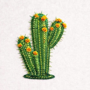 Op de afbeelding: Geborduurd cactusontwerp in groen- en geeltinten, met kleine oranje bloemen. De cactus heeft een gedetailleerd, getextureerd uiterlijk en staat tegen een witte achtergrond. De borduursel is waarschijnlijk voor een patch of applicatie.