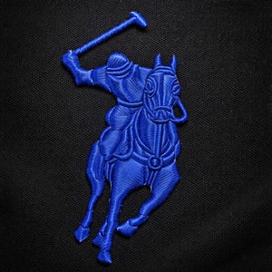 May include: A blue embroidered patch featuring a polo player on a horse, holding a mallet. The design is in a detailed, textured style, with the horse and rider in a dynamic pose. The patch is set against a black background.