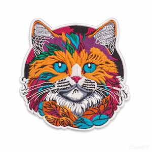 May include: Embroidered patch of a cat's face in vibrant colours. The cat has orange, purple, and teal fur, blue eyes, and white whiskers. The patch is framed by a black border and white stitching.