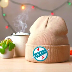 Puede incluir: Un gorro beige con un parche circular con un diseño turquesa y rojo. El gorro está junto a una taza blanca con una bebida caliente y una pequeña planta suculenta en una maceta blanca. Luces decorativas en el fondo.