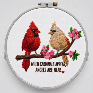Op de afbeelding: Geborduurde hoepelkunst met twee kardinalen op een tak met roze bloesems. Eén kardinaal is rood, de andere is bruin. De tekst luidt: "WHEN CARDINALS APPEAR, ANGELS ARE NEAR" met een rood hart.