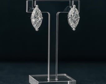 May include: A pair of marquise-cut diamond earrings on a clear acrylic display stand. The earrings are set in a silver metal and have a brilliant sparkle. The stand has a square base and a T-shaped top.