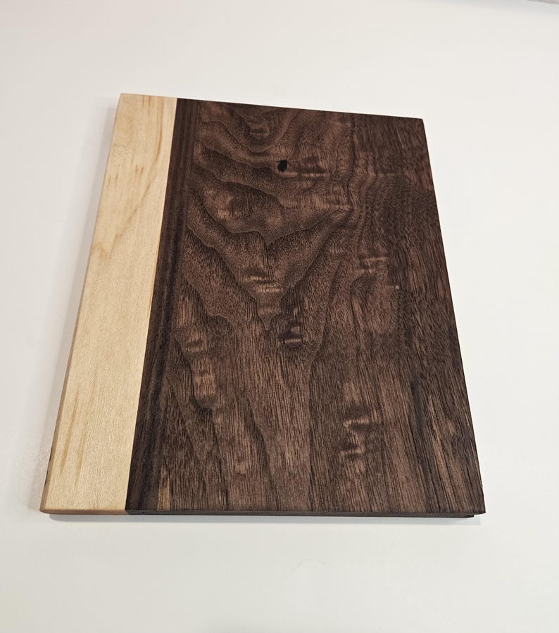 May include: A rectangular wooden cutting board with a light-colored wood section on the left and a dark brown wood section on the right. The wood grain is visible, and the board is set against a white background.