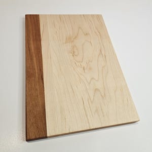 May include: A rectangular wooden cutting board with a light-colored wood grain surface and a dark brown wood border on the left side. The board is made of natural wood and has a smooth finish.