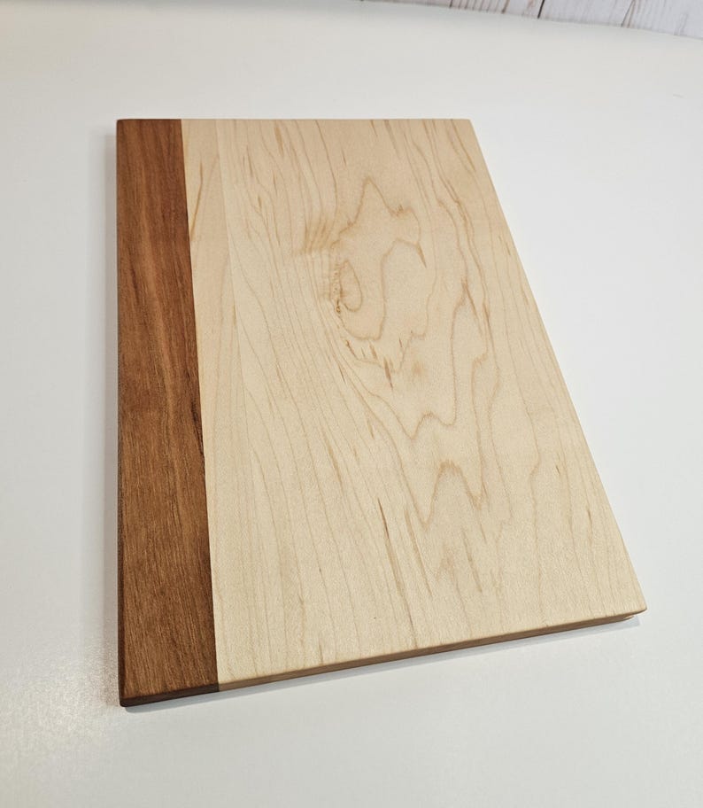May include: A rectangular wooden cutting board with a light-colored wood grain surface and a dark brown wood border on the left side. The board is made of natural wood and has a smooth finish.