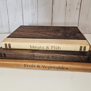 May include: Three wooden cutting boards stacked on top of each other. The top board is dark brown, the middle board is light brown, and the bottom board is a lighter shade of brown. Each board has text: "Meats & Fish", "Cheese & Bread", and "Fruit & Vegetables".