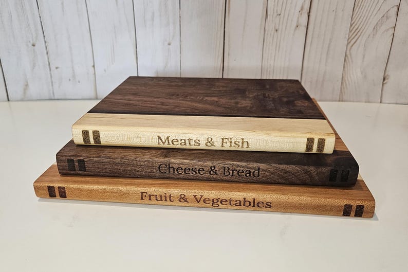 May include: Three wooden cutting boards stacked on top of each other. The top board is dark brown, the middle board is light brown, and the bottom board is a lighter shade of brown. Each board has text: "Meats & Fish", "Cheese & Bread", and "Fruit & Vegetables".