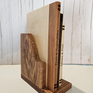 May include: A wooden bookend with a dark brown base and a light brown back. The bookend holds three wooden cutting boards with the words "Fruit & Vegetables", "Cheese & Bread", and "Meats & Fish" engraved on them. The bookend is made of various wood tones.