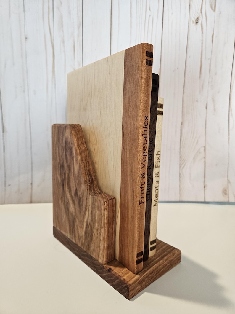 May include: A wooden bookend with a dark brown base and a light brown back. The bookend holds three wooden cutting boards with the words "Fruit & Vegetables", "Cheese & Bread", and "Meats & Fish" engraved on them. The bookend is made of various wood tones.