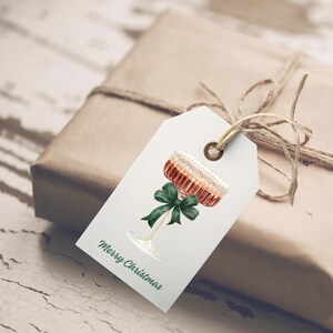 Op de afbeelding: Een cadeaulabel met een aquarel illustratie van een cocktail met een groene strik. Het label is bevestigd aan een ingepakt cadeau met touw. De woorden "Merry Christmas" staan op het label.