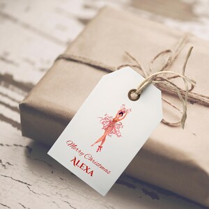 Könnte beinhalten: Ein weißes Geschenk-Etikett mit einer roten Ballerina-Illustration und dem Text "Merry Christmas ALEXA". Das Etikett ist mit einer Schnur an einem verpackten Geschenk befestigt. Die Ballerina trägt einen roten Tutu.