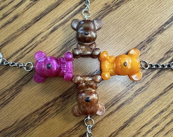 May include: A collection of four bear-shaped keychains in shades of brown, orange, and pink, connected by silver chains and rings. The keychains are arranged in a cross shape, with a rose illustration in the upper left corner.