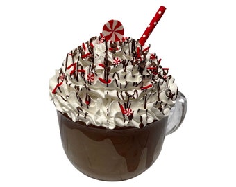May include: A white ceramic mug filled with a chocolate drink topped with whipped cream, chocolate sauce, red and white candy sprinkles, and a red and white candy cane. A red and white striped straw sticks out of the top.