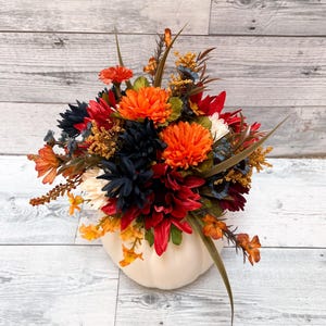 May include: A fall-themed floral arrangement in a white pumpkin-shaped vase. The arrangement features orange, red, navy, and yellow flowers with green foliage. The vase is made of ceramic or a similar material.