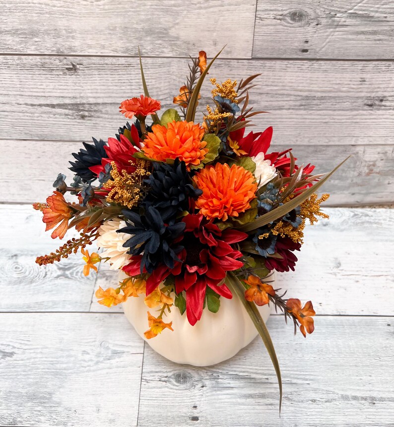 May include: A fall-themed floral arrangement in a white pumpkin-shaped vase. The arrangement features orange, red, navy, and yellow flowers with green foliage. The vase is made of ceramic or a similar material.