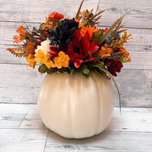 May include: A cream-colored pumpkin-shaped vase filled with a vibrant arrangement of fall-colored flowers. The flowers include orange, red, yellow, and dark blue blooms, with green foliage and tall, slender grass accents. The vase sits on a light wood-grain surface.