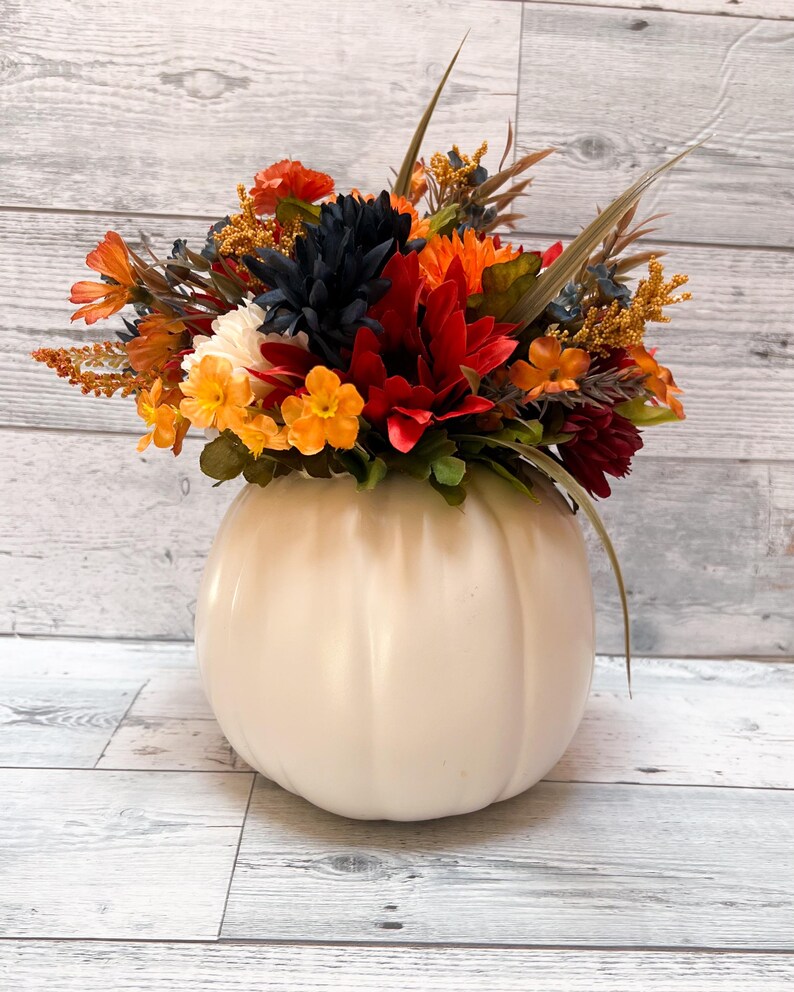 May include: A cream-colored pumpkin-shaped vase filled with a vibrant arrangement of fall-colored flowers. The flowers include orange, red, yellow, and dark blue blooms, with green foliage and tall, slender grass accents. The vase sits on a light wood-grain surface.