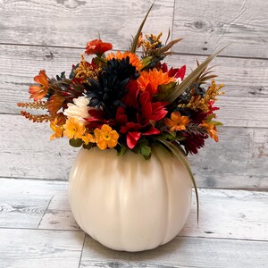 May include: A cream-colored pumpkin-shaped vase filled with a vibrant arrangement of fall flowers. The flowers include orange, red, yellow, and dark blue blooms, with green foliage and brown accents. The vase sits on a light wood-grain surface.