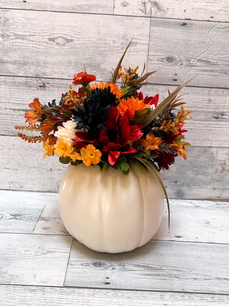May include: A cream-colored pumpkin-shaped vase filled with a vibrant arrangement of fall flowers. The flowers include orange, red, yellow, and dark blue blooms, with green foliage and brown accents. The vase sits on a light wood-grain surface.