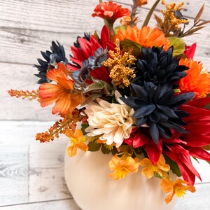 May include: A fall-themed floral arrangement in a cream-colored pumpkin-shaped vase. The arrangement features a variety of colorful flowers in shades of red, orange, navy, and cream. The background is a light-colored wooden surface.