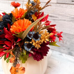 May include: A cream-colored pumpkin-shaped vase filled with a vibrant arrangement of artificial flowers. The flowers include orange, red, navy blue, and yellow blooms, with green foliage. The arrangement is set against a light wood-grain background.