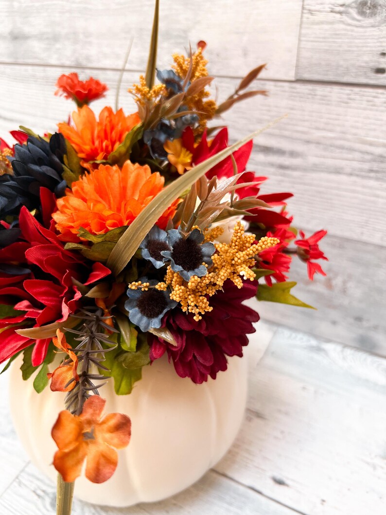 May include: A cream-colored pumpkin-shaped vase filled with a vibrant arrangement of artificial flowers. The flowers include orange, red, navy blue, and yellow blooms, with green foliage. The arrangement is set against a light wood-grain background.