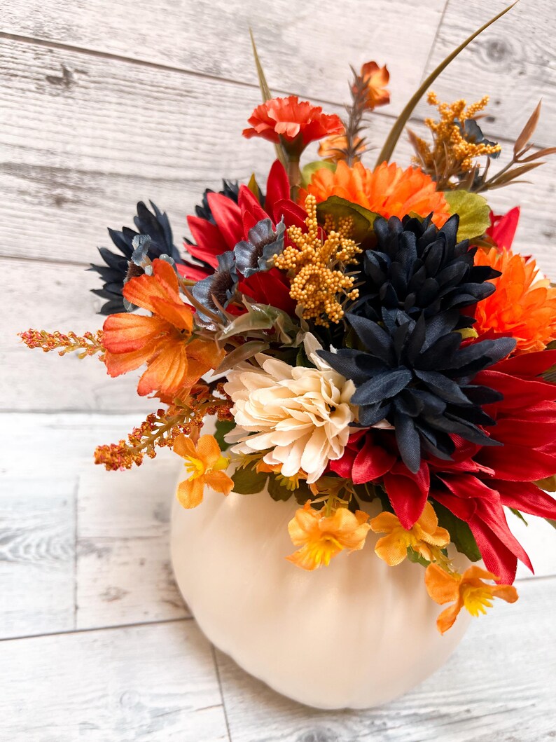 May include: A fall-themed floral arrangement in a cream-colored pumpkin-shaped vase. The arrangement features a variety of colorful flowers in shades of red, orange, navy, and cream. The background is a light-colored wooden surface.