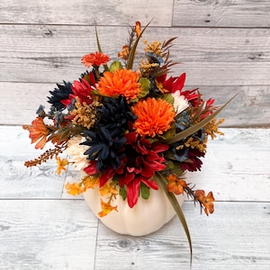 May include: A fall-themed floral arrangement in a cream-colored pumpkin-shaped vase. The arrangement features orange, red, navy, and white artificial flowers and greenery. The pumpkin vase is approximately 8 inches wide.