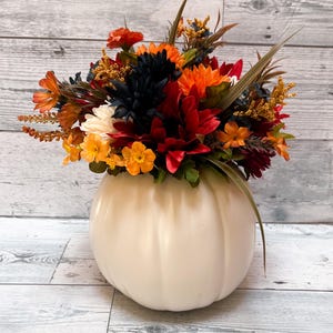 May include: A cream-colored pumpkin-shaped vase filled with a vibrant arrangement of fall-colored flowers. The flowers include orange, red, yellow, and dark blue blooms, with green and brown accents. The vase is sitting on a light wood-grain surface.