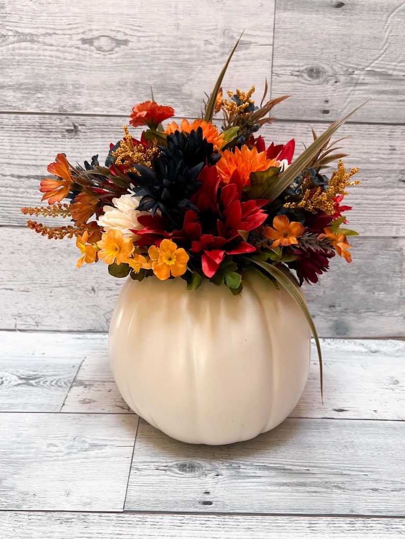 May include: A cream-colored pumpkin-shaped vase filled with a vibrant arrangement of fall-colored flowers. The flowers include orange, red, yellow, and dark blue blooms, with green and brown accents. The vase is sitting on a light wood-grain surface.
