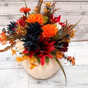 May include: A fall-themed floral arrangement in a cream-colored pumpkin-shaped vase. The arrangement features orange, red, navy, and yellow flowers with green and brown accents. The vase sits on a white wooden surface.