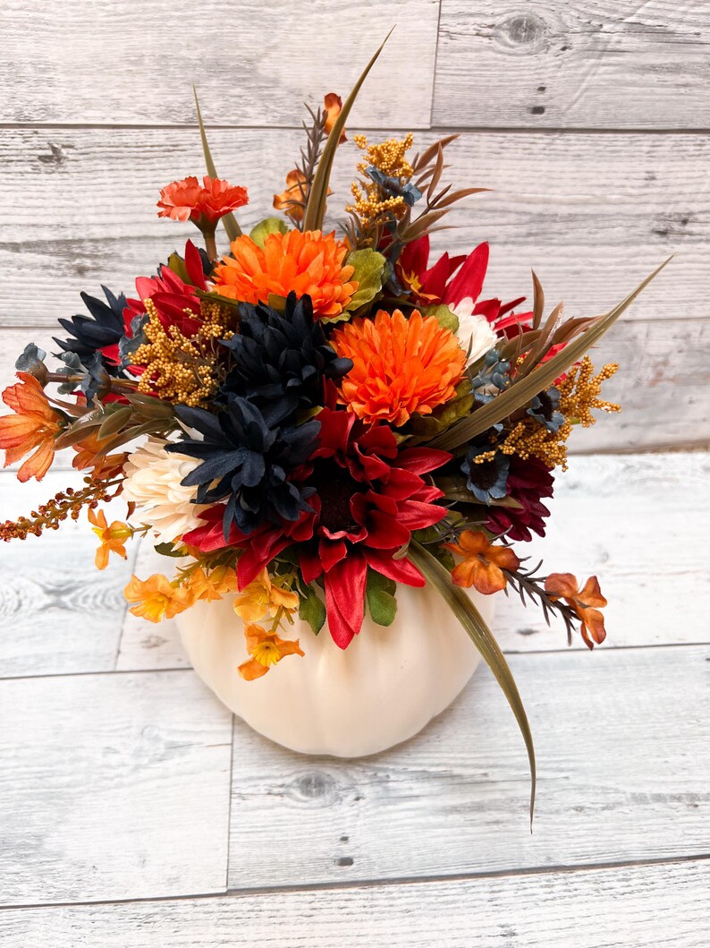May include: A fall-themed floral arrangement in a cream-colored pumpkin-shaped vase. The arrangement features orange, red, navy, and yellow flowers with green and brown accents. The vase sits on a white wooden surface.