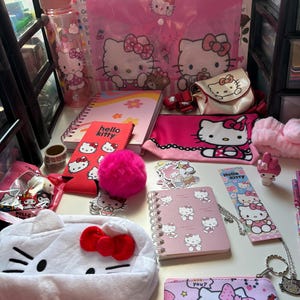 May include: A selection of pink Hello Kitty themed items, including a water bottle, notebooks, a pencil case, stickers, and a coin purse. The items are displayed on a white surface, with "Hello Kitty" text on several items.