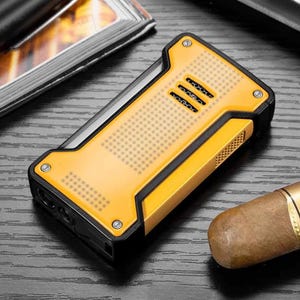 May include: A yellow and black rectangular lighter with a textured surface and three horizontal vents. The lighter has a metallic silver trim and is shown next to a cigar on a dark wooden surface.